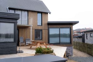 Façade contemporaine signée WoodUpp : bardage à lames verticales en bois naturel sur la maison et large terrasse en lames de bois WoodUpp, avec grande baie vitrée coulissante noire, finitions anthracite autour des fenêtres et jardinière posée sur la terrasse.