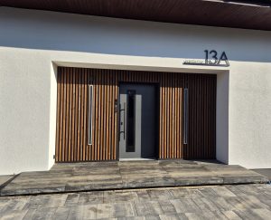 Entrée moderne d'une maison présentant un bardage WoodUpp à lames verticales en bois naturel encadrant une porte grise avec poignée noire, deux éclairages verticaux et le numéro 13A. Devant, une terrasse WoodUpp en dalles boisées sur dallage gris complète l'aménagement minimaliste.