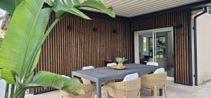 Terrasse couverte avec bardage vertical WoodUpp composé de lames de bois foncé aux finitions foncées, espacées et cadre métallique noir, protégeant une baie vitrée. Mobilier extérieur: grande table grise et chaises en rotin, plante verte en premier plan. Ambiance contemporaine et cosy.