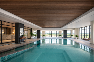 Une piscine intérieure moderne et luxueuse avec une rampe d'accès et de grandes fenêtres. Le plafond est magnifiquement habillé de panneaux acoustiques en tasseaux de bois WoodUpp. Ces panneaux WoodUpp, dans une finition bois foncé, ajoutent de la chaleur et une texture linéaire élégante au-dessus de l'eau turquoise.