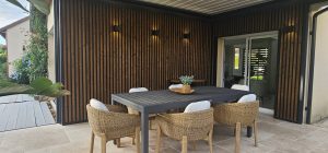 Terrasse couverte montrant un bardage WoodUpp à lames verticales en bois foncé avec cadre noir et appliques murales, une table noire moderne entourée de fauteuils en osier, et, sur la gauche, un platelage WoodUpp gris à lames larges, créant un espace extérieur chaleureux et contemporain.