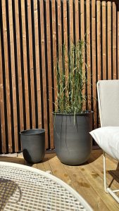 Plan rapproché d'une terrasse WoodUpp : brise-vue vertical en lames de bois naturel WoodUpp, teinte chaude, plancher de terrasse en lames de bois WoodUpp, deux pots gris (un grand contenant des herbes hautes, un petit vide), chaise et table blanches partiellement visibles au soleil.