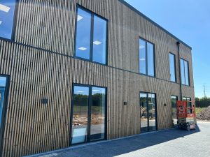 Façade de bâtiment WoodUpp revêtue d'un bardage à lames de bois verticales WoodUpp, finition naturelle et texture striée, sur deux étages, encadrant plusieurs grandes baies vitrées et portes-fenêtres à menuiseries sombres. Au sol, pavés et une nacelle élévatrice rouge, ciel bleu en arrière-plan.