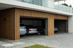Façade de garage moderne habillée par WoodUpp : bardage à lames verticales en bois clair, lattes fines et espacées encadrant trois portes de box ouvertes, contraste avec profilés noirs. On devine trois voitures garées à l’intérieur, sol en dalles grises et balcon vitré au‑dessus.