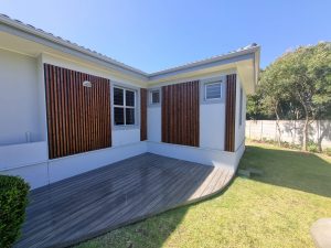 Façade extérieure d'une maison équipée par WoodUpp : grands panneaux de lamelles de bois verticales brun foncé installés autour des fenêtres et une terrasse en lames composite gris WoodUpp à bord arrondi, soignée et moderne. Jardinet et haie complètent l'aménagement extérieur ensoleillé.