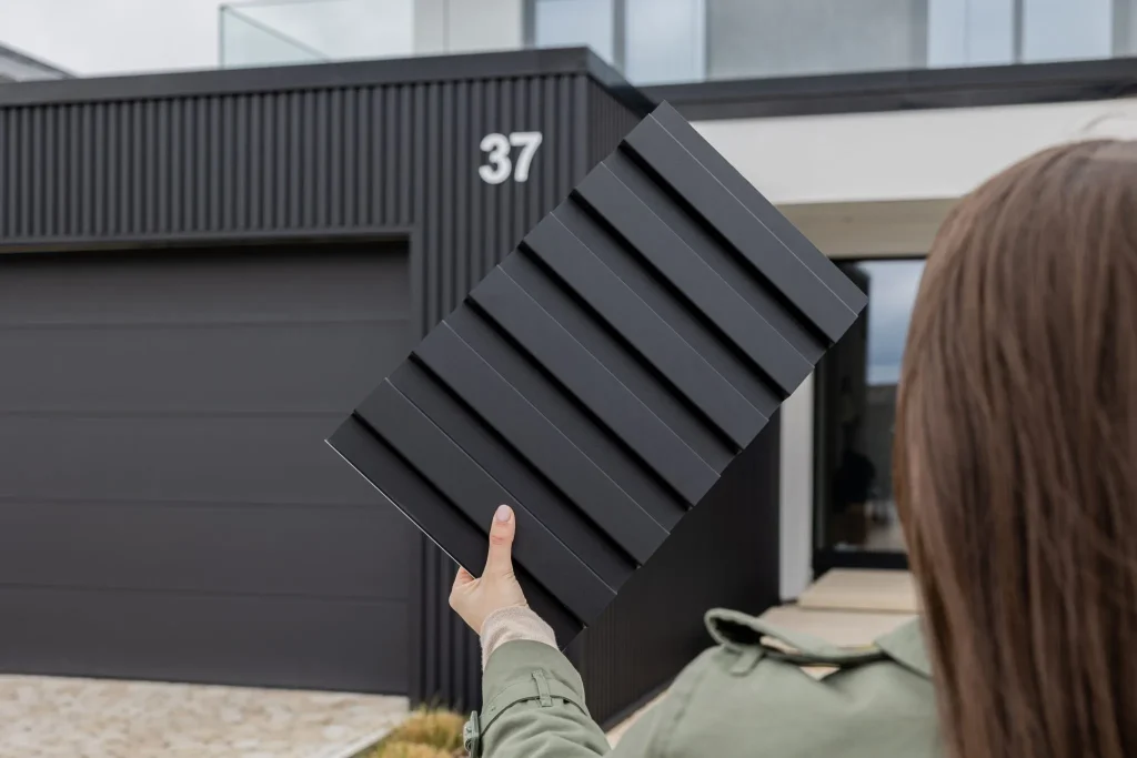 Vue d’une main tenant un échantillon rectangulaire de panneau WoodUpp noir anthracite à finition mate, nervures régulières en relief (bardage profilé), devant une maison moderne revêtue du même bardage WoodUpp à lames verticales noires, façade minimaliste, avec un grand garage assorti.