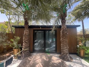 Façade d'une annexe de jardin WoodUpp avec bardage à lames verticales en bois naturel, encadrement noir et double porte vitrée, entourée de deux palmiers et pots dorés; la finition boisée WoodUpp crée un mur ajouré élégant filtrant la lumière estivale.