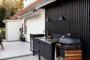Coin cuisine extérieur WoodUpp : meuble modulaire noir avec surface inox et évier intégré, portes de rangement, petit réfrigérateur encastré, deux suspensions noires et un grand barbecue kamado muni d'une tablette latérale en bois WoodUpp, posé devant un mur à lames verticales noires.
