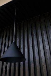 Mur lambrissé WoodUpp en lames verticales de bois brûlé noir, finition rainurée, accompagné d'une suspension conique noire descendante. Gros plan montrant la texture des veines du bois, les fixations apparentes et le contraste sobre entre la lampe et le bardage WoodUpp.
