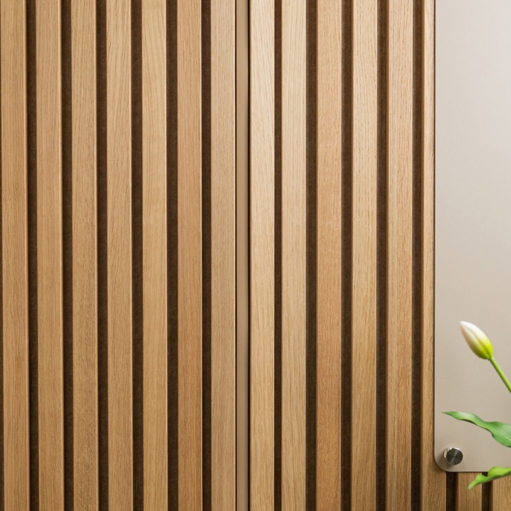 Panneau mural WoodUpp à lames verticales en chêne naturel, panneau acoustique aux rainures sombres et finition huilée, couvrant la majeure partie de l'image. À droite, on aperçoit un panneau clair fixé par une entretoise métallique WoodUpp et le bouton floral d'une tulipe.