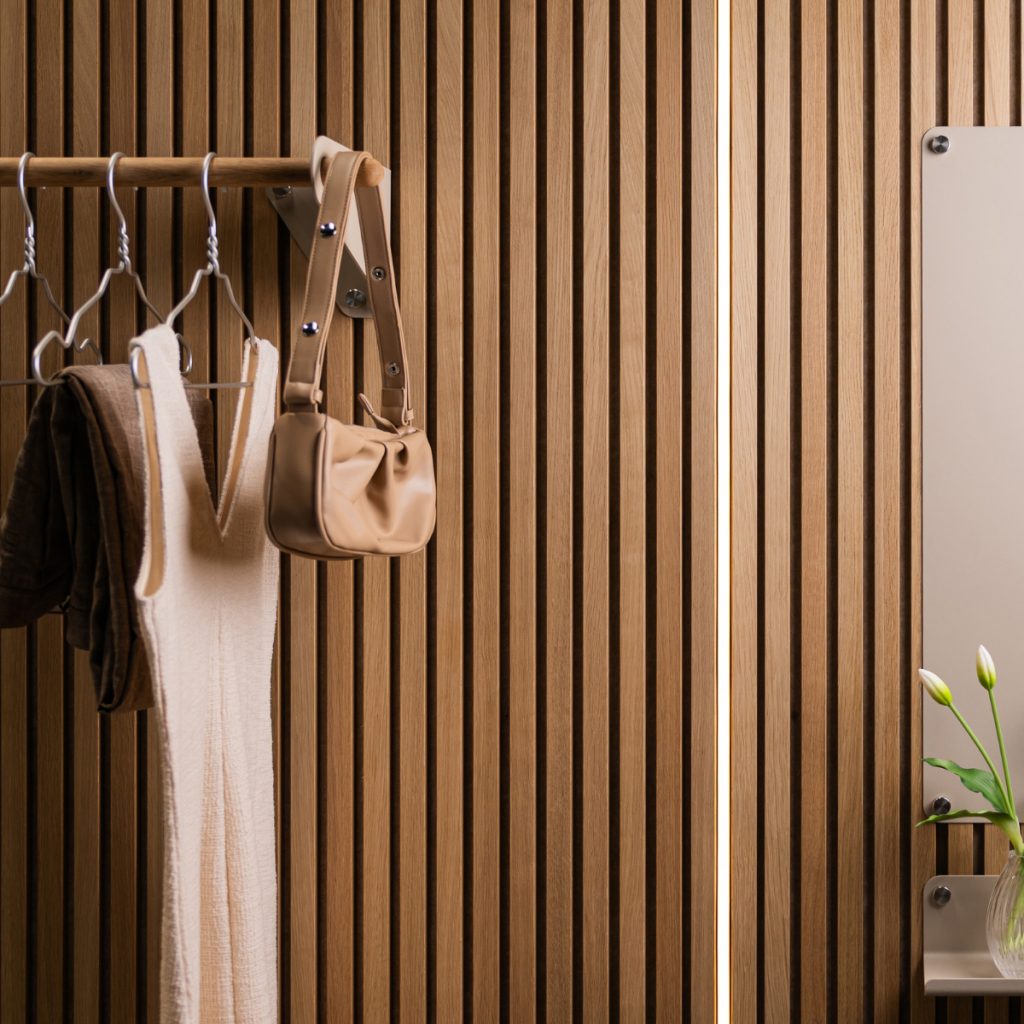 Mur à lattes en chêne WoodUpp, finition naturelle, accompagné d'une barre porte-manteaux WoodUpp en bois avec cintres métalliques, sac et vêtements suspendus; à droite, miroir et petite tablette WoodUpp en métal clair avec vase de tulipes, éclairage vertical discret, élégant.