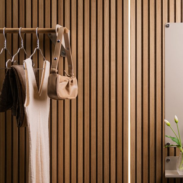 Mur à lattes en chêne WoodUpp, finition naturelle, accompagné d'une barre porte-manteaux WoodUpp en bois avec cintres métalliques, sac et vêtements suspendus; à droite, miroir et petite tablette WoodUpp en métal clair avec vase de tulipes, éclairage vertical discret, élégant.