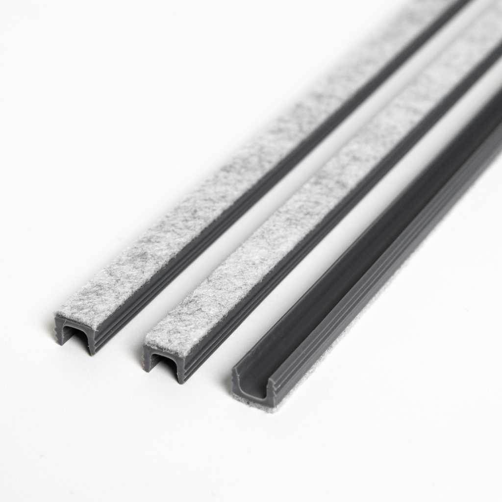 Photo montrant pour WoodUpp trois profilés en U en plastique gris anthracite, alignés, avec une bande de feutre gris clair collée sur leur face supérieure. Produits WoodUpp destinés à protection et étanchéité des bords : rails profilés étroits, finition texturée, angles ouverts visibles.