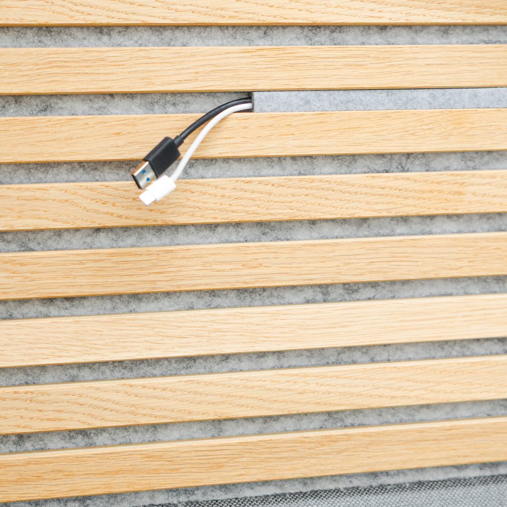 Panneau acoustique WoodUpp à lattes de chêne clair, espacées, avec feutre gris en arrière-plan; encoche de passage de câbles intégrée laissant sortir deux câbles (USB et chargeur). Finition mate et grain du bois visibles, design mural décoratif et acoustique élégant.