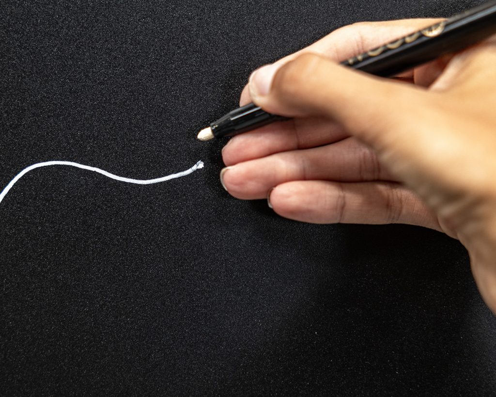 Gros plan WoodUpp montrant une main traçant une ligne blanche avec un feutre craie liquide blanc WoodUpp au corps noir et embout biseauté, sur un tableau ardoise noir WoodUpp à surface mate légèrement granuleuse. La trace est sinueuse et nette, illustrant précision et opacité du marqueur.