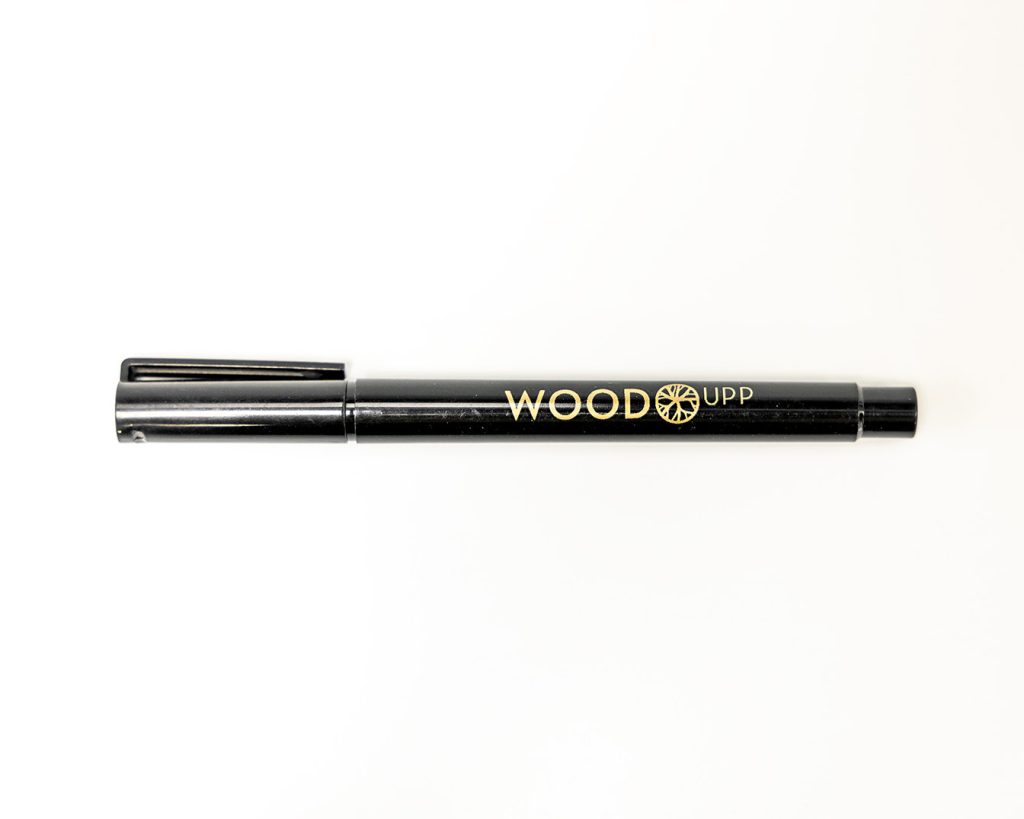 Feutre noir WoodUpp posé horizontalement sur fond blanc, capuchon à gauche avec clip. Corps cylindrique brillant orné du logo doré «WOODUPP» et d’un pictogramme circulaire stylisé. Embout droit apparent, surface lisse et aspect de marqueur fin destiné à l’écriture ou au marquage.