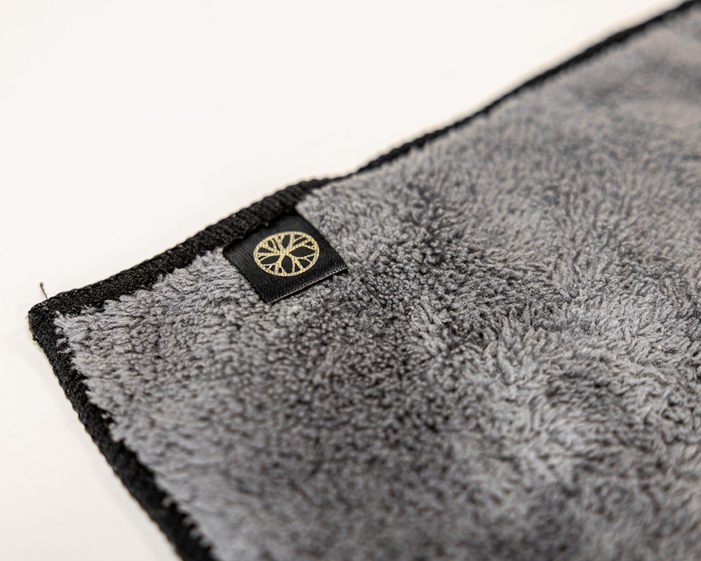 Plan rapproché d'un chiffon microfibre gris WoodUpp, texture pelucheuse et dense, bordure noire cousue et étiquette noire portant le logo doré de WoodUpp en forme de cercle stylisé. Produit présenté sur fond clair, mettant en valeur la qualité et la finition du tissu.