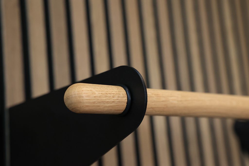 Image montrant une barre en chêne massif WoodUpp à embout arrondi, insérée dans un support mural circulaire noir WoodUpp, fixée sur une planche à lames verticales. La finition bois claire et le métal noir mat créent un contraste moderne et épuré.