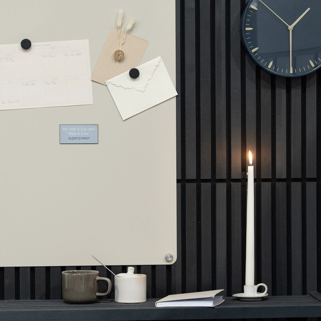 Coin de bureau avec tableau magnétique WoodUpp crème, fixé par entretoises métalliques, décoré de notes et aimants, posé devant des panneaux muraux WoodUpp à lames de bois noires. Sur la tablette: tasse, sucrier, carnet et une bougie allumée à côté d'une horloge murale.