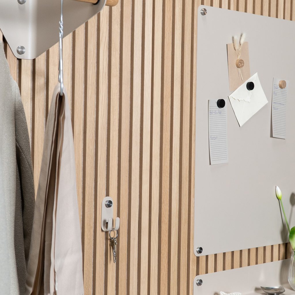 Panneau mural WoodUpp à lames verticales en chêne clair, avec crochets métalliques blancs WoodUpp tenant clés et vêtements, et plaques magnétiques WoodUpp beige fixées par caches métalliques. Notes et aimants ronds noirs punaisés sur la plaque; vase tulipe en verre posé en bas à droite.