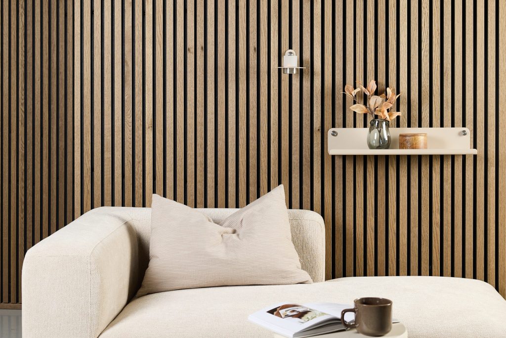 Image d'un salon minimaliste avec un mur WoodUpp en lamelles verticales à rainures fines en chêne clair et noir, et une étagère murale WoodUpp beige fixée par vis apparentes; canapé crème, coussin, vase décoratif, bougie et tasse posés sur une table basse.