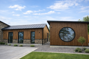 Vue extérieure de deux bâtiments modernes utilisant le bardage extérieur vertical de WoodUpp. Le bardage en bois, aux tons chauds et riches, couvre entièrement la façade de droite avec sa grande fenêtre ronde, et la partie supérieure du bâtiment de gauche au-dessus d'un soubassement en pierre.
