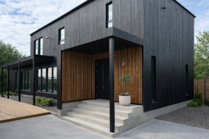 Extérieur d'une maison moderne au bardage noir, où l'entrée est sublimée par des panneaux acoustiques WoodUpp. Ces panneaux muraux présentent des tasseaux de bois naturel aux tons chauds sur un fond de feutre noir, apportant une texture riche et un contraste élégant à l'architecture sombre.