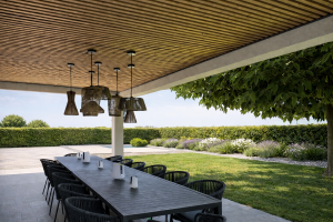 Une terrasse couverte moderne présente un plafond habillé de panneaux acoustiques en tasseaux de bois clair WoodUpp. Une longue table sombre entourée de chaises noires et de luminaires suspendus en osier surplombe un jardin soigneusement entretenu et une piscine.