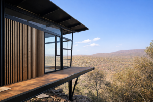 Cette image montre une architecture moderne intégrée à la nature. On y voit un bardage extérieur en tasseaux de bois de WoodUpp sur la gauche, apportant chaleur et texture à la structure en acier noir et à la terrasse en bois face à la vue panoramique.