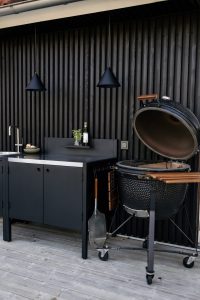 Coin barbecue WoodUpp : meuble de cuisine d'extérieur noir WoodUpp avec évier et plan de travail, petite étagère murale contenant une bouteille et des herbes, deux suspensions coniques noires, barbecue kamado noir à couvercle ouvert avec poignée et tablette latérale en bois, terrasse en lames grises.