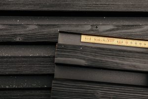 Image montrant des lames de bardage WoodUpp noires, texturées avec grain apparent et légers résidus blancs, posées horizontalement; un mètre pliant en bois glissé entre deux lames indique l'échelle. Produit WoodUpp : lames de façade noires à finition bois, rainurées et modernes.